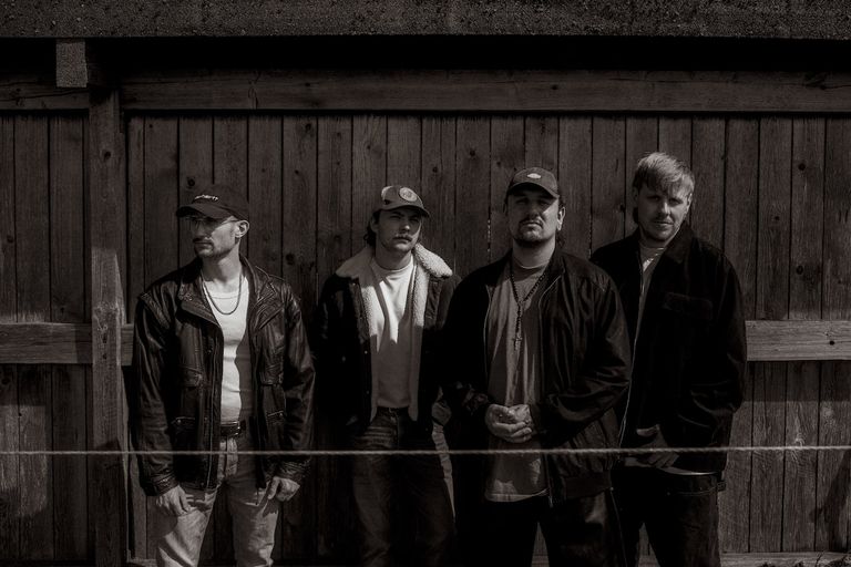 Heavy music band Half Me stands together in dark jackets and baseball caps against a rustic wooden fence backdrop in a moody promo photo.